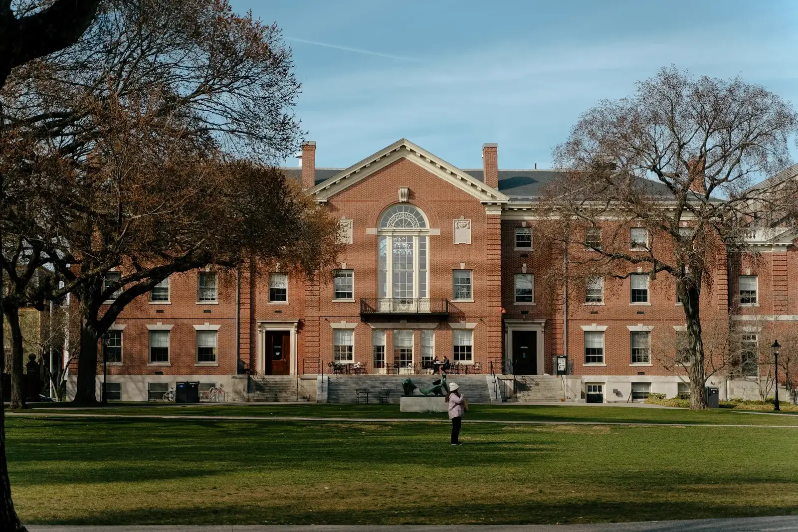 A red brick university hall behind a lawn and bare winter trees, one person standing in front of it.