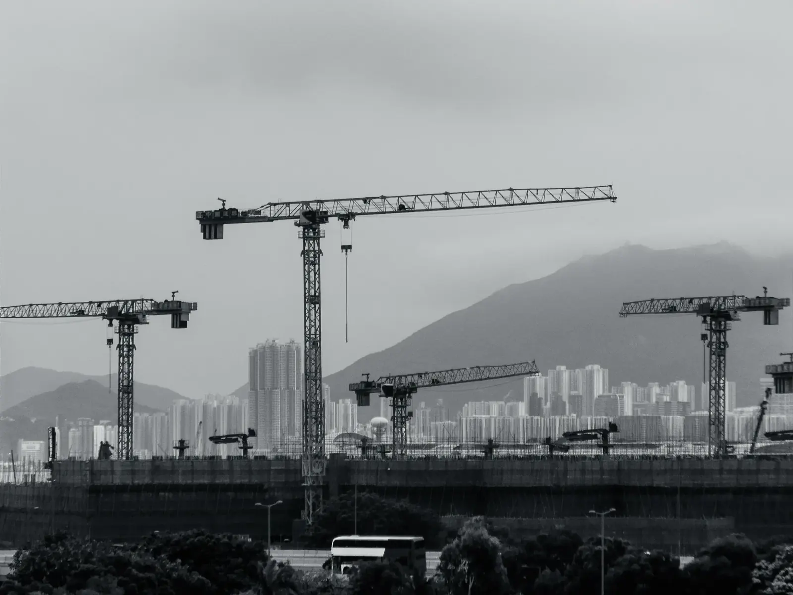 Tower cranes above a construction site in black and white, a mountain and a dense city skyline behind them.