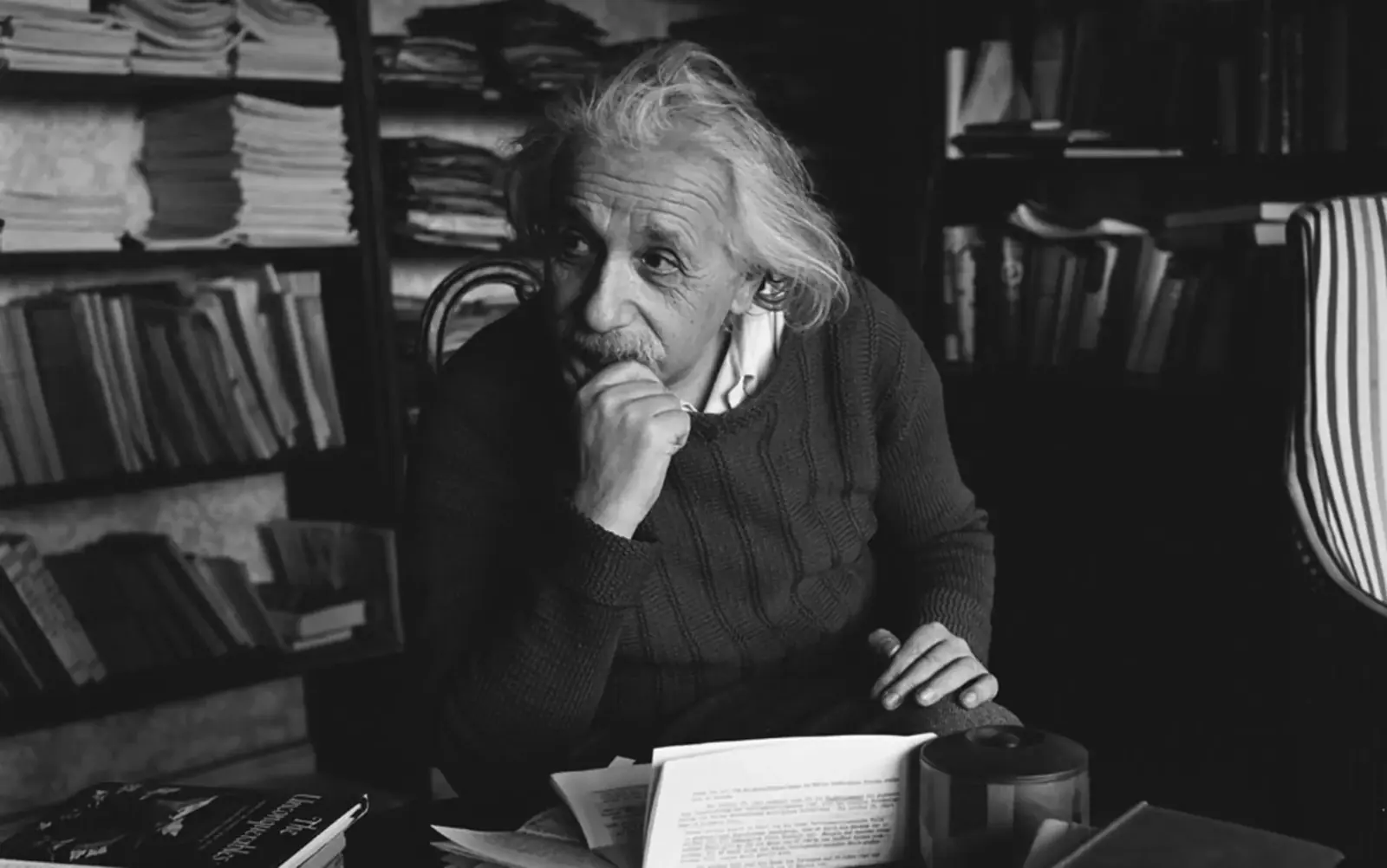 Albert Einstein sitting at a paper-strewn desk in his study, chin resting on his hand, shelves of books and journals behind him.
