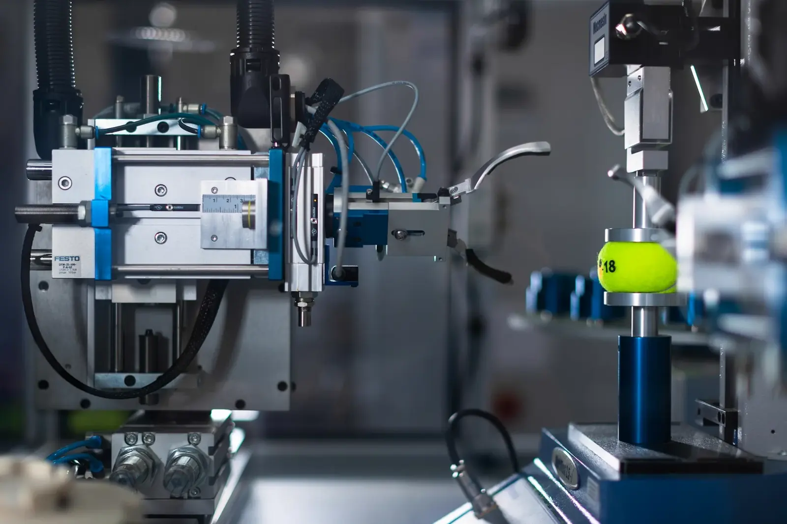 A pneumatic robotic arm on an automated test rig, closing on a tennis ball held on a spindle.