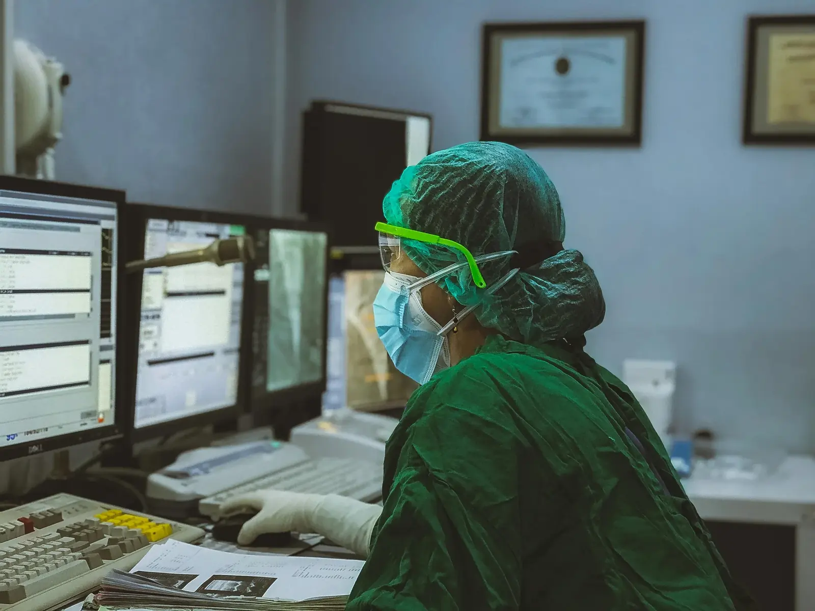 A clinician in green scrubs, cap and mask working at a bank of monitors showing medical scans.